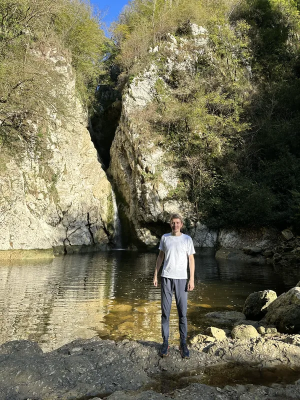 Агурские водопады: турист стоит на камнях у спокойной воды перед чашей водопада, тонкая струя падает из щели между двумя светлыми скальными стенами