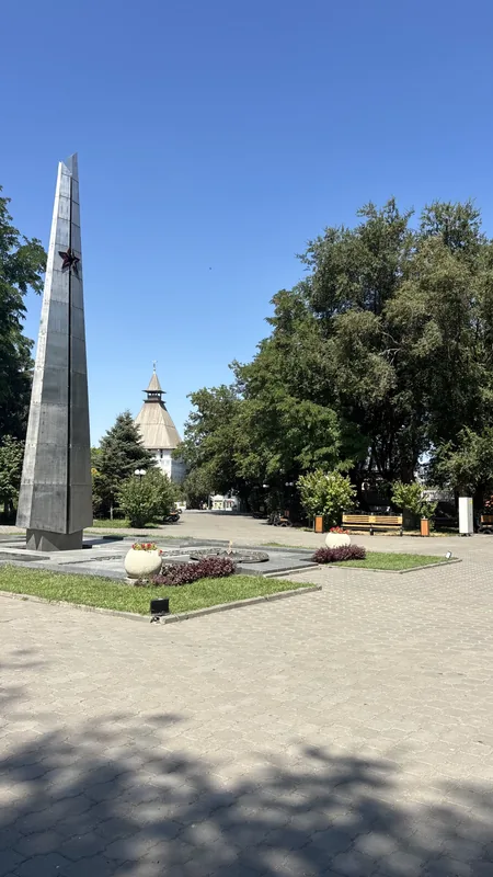 Братский сад: гранитный обелиск со звездой на клумбе, за ним видна одна из башен кремля и аллея с газоном.