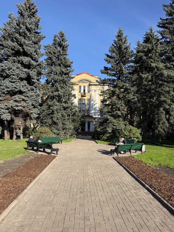 Аллея парка с голубыми елями и зелёными скамейками, в конце — жёлтое здание с колоннами
