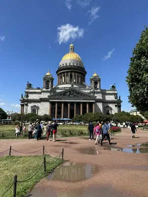 Исаакиевский собор с золотым куполом в ясный день, перед ним газоны, туристы и лужи на дорожке после дождя