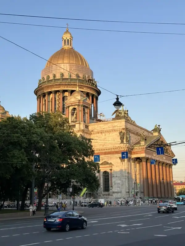 Собор в вечернем солнце с угла площади: у барабана колоннады видны строительные конструкции, впереди дорога с машинами