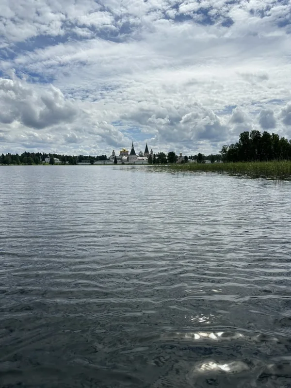 Иверский монастырь с воды: белые стены и главы собора узкой полосой на дальнем берегу, широкая гладь Валдайского озера и облачное небо