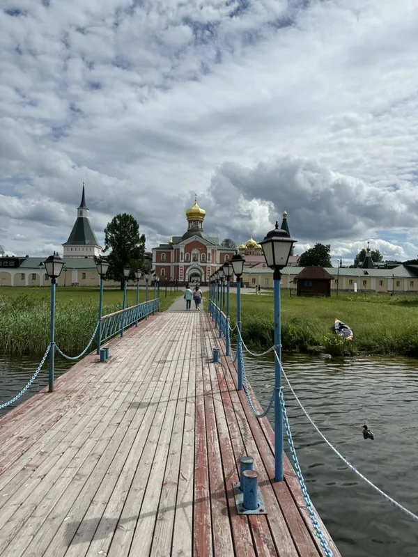 Деревянный настил-пирс над водой: голубые фонари и цепи вдоль перил, впереди краснокирпичный Иверский собор с золотыми куполами и белая колокольня с зелёным шатром