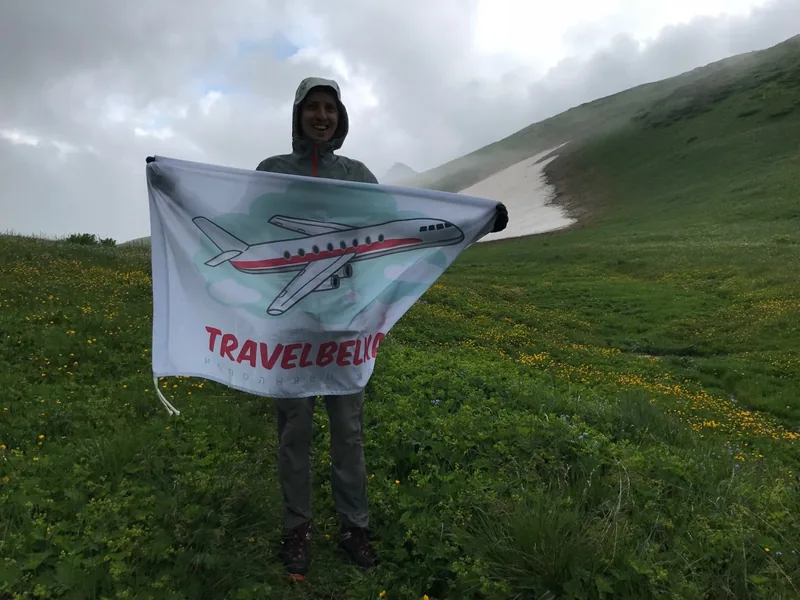 Кавказский заповедник: турист с флагом TravelBelka на горном склоне у пятна снега, туман и низкие облака.