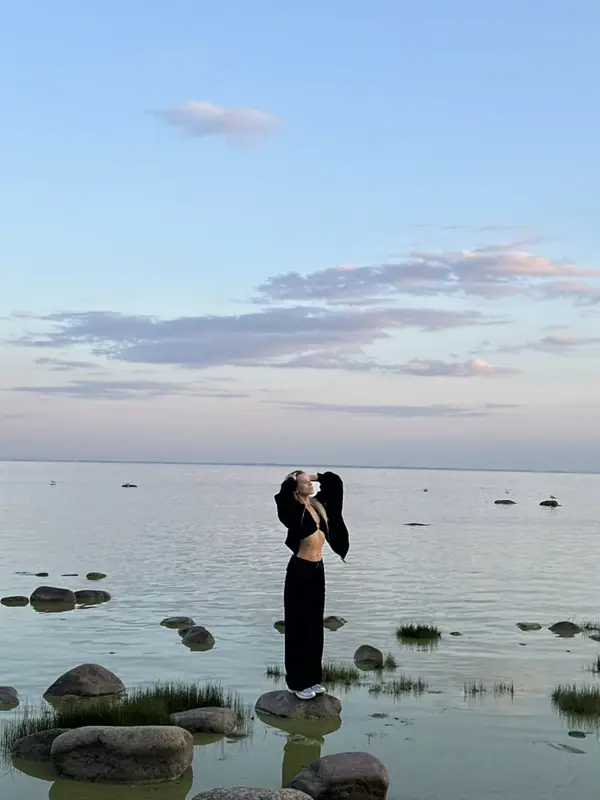 Девушка в чёрном стоит на валуне в мелкой воде среди камней и травяных кочек, вечернее небо с розовыми облаками