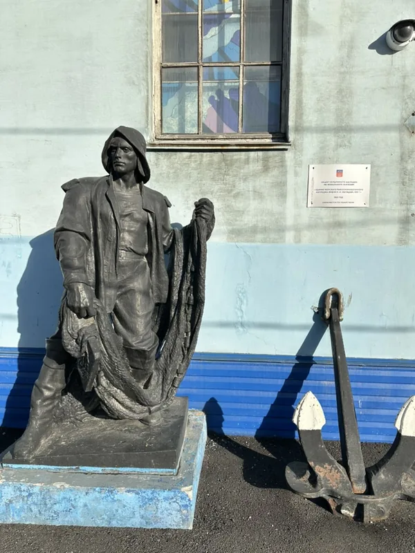 Бронзовая фигура рыбака в зюйдвестке и робе с сетью в руках у голубой стены в Мурманске, рядом на асфальте лежит старый корабельный якорь