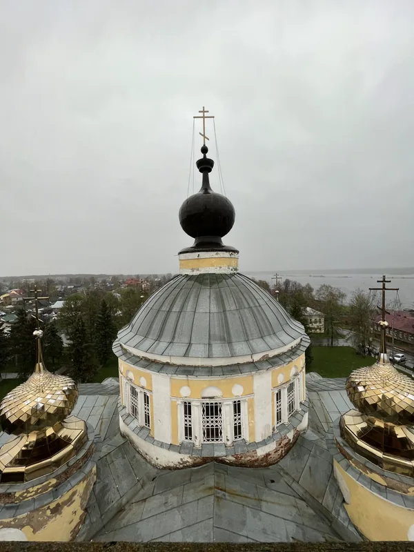 Вид с колокольни на купол собора: серая кровля и чёрная главка с крестом, по краям кадра золотые главки, внизу крыши города и река