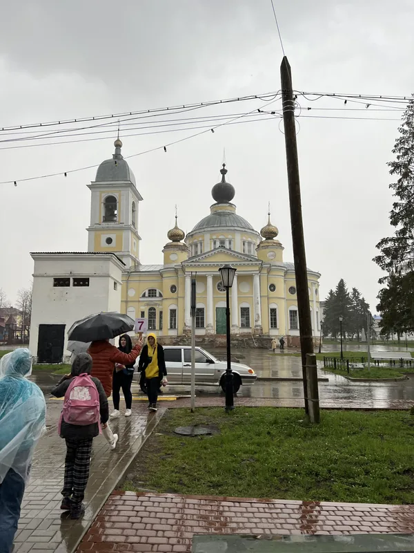 Жёлтый собор с золотыми главками и белой колокольней в дождливый день, по мокрому тротуару идут люди в дождевиках и с зонтами