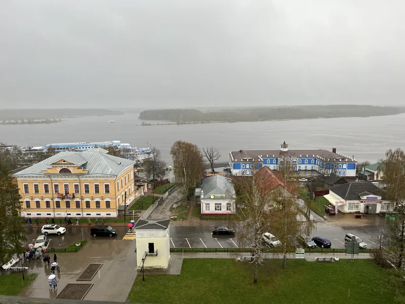 Вид сверху на приволжский городок: жёлтый двухэтажный особняк, синие дома у пристани, за ними широкая река в серой дымке