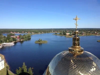 Золотой купол с крестом крупным планом, за ним синее озеро с лесистыми островами, у дальнего берега стоит белый теплоход