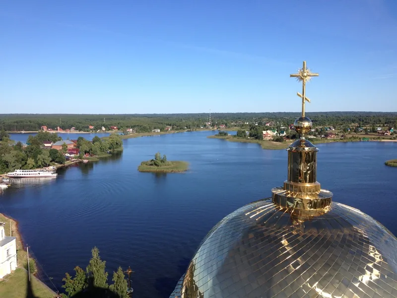 Золотой купол с крестом крупным планом, за ним синее озеро с лесистыми островами, у дальнего берега стоит белый теплоход