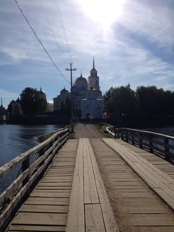 Деревянный настил-мост через воду ведёт к белому монастырю с колокольней и куполами, солнце светит прямо в объектив