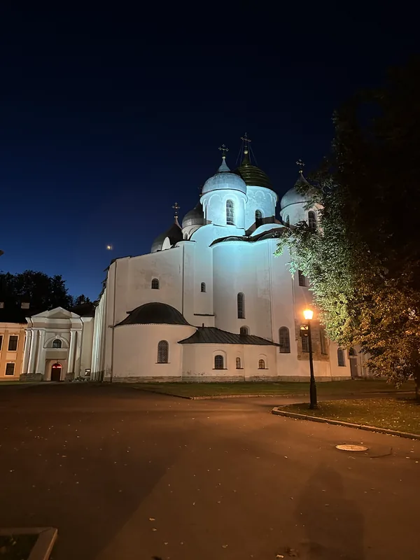 Софийский собор в вечерней подсветке: белые стены, шлемовидные купола, золотой центральный, рядом фонарь на пустой дорожке