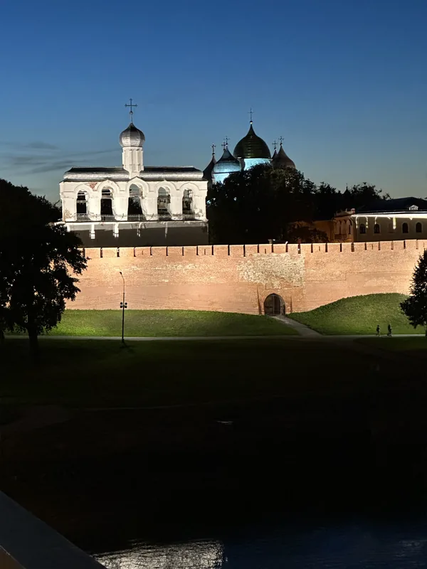 Новгородский детинец вечером: подсвеченная краснокирпичная стена на валу, за ней звонница с арками и золотые купола Софийского собора