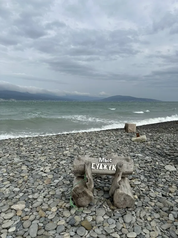 Новороссийск: деревянная табличка «Мыс Суджук» на галечном пляже, штормовое море и облачное небо.
