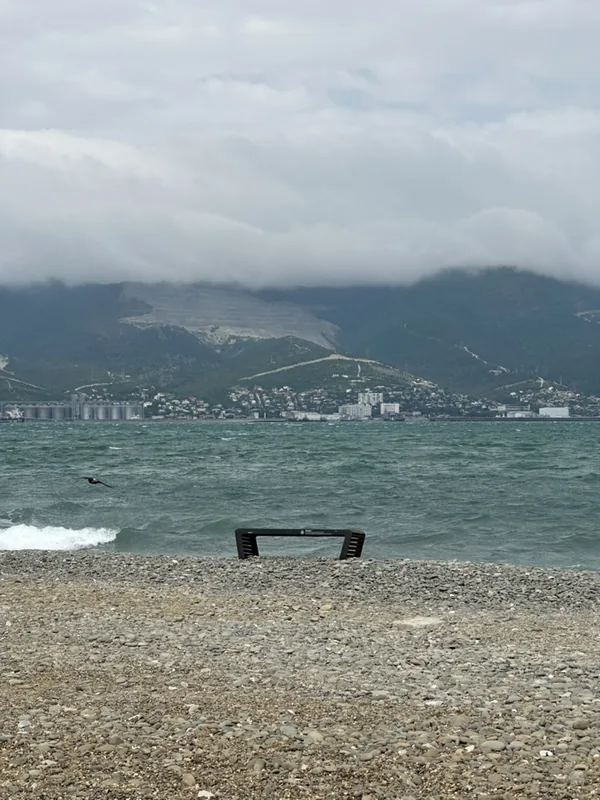 Новороссийск: галечный пляж со скамейкой у воды, вид на бухту, город и горы в облаках.