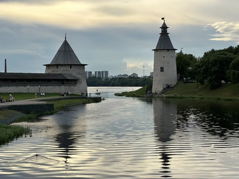 Плоская и Высокая башни у слияния Псковы и Великой в вечернем свете, на берегу под стенами гуляют люди