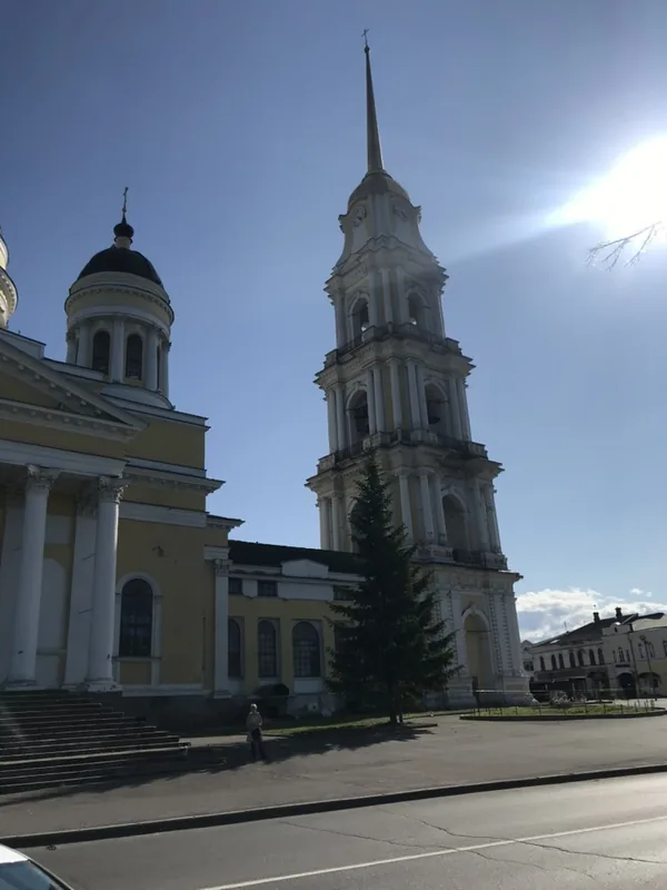 Высокая ярусная колокольня со шпилем рядом с собором, перед ними ель, солнце светит из-за колокольни