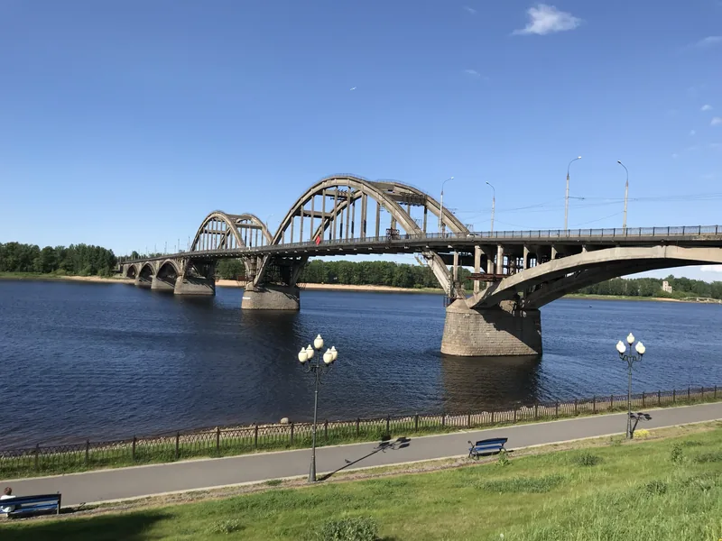 Арочный мост через широкую реку, на переднем плане набережная с фонарями и скамейками на зелёном газоне