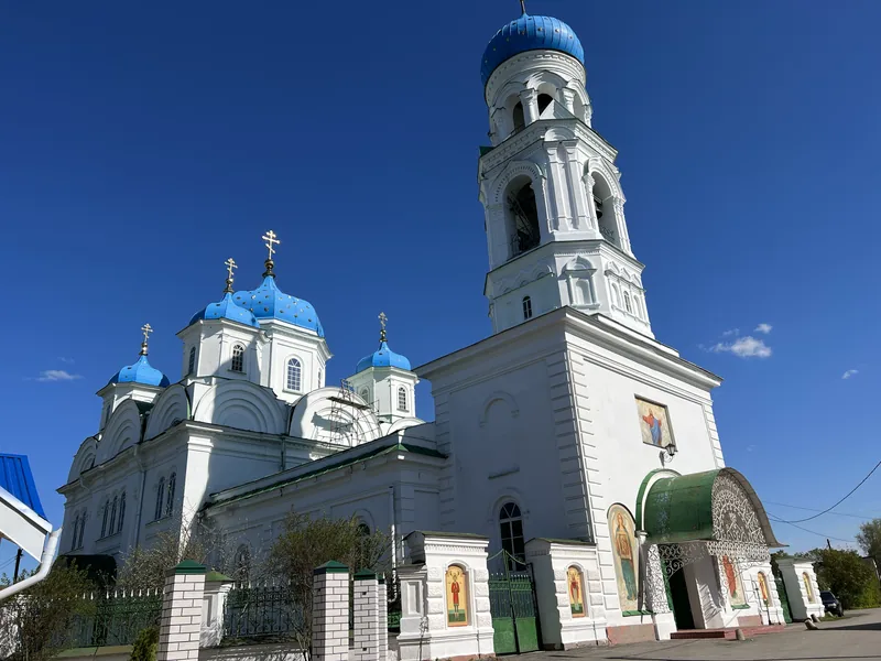 Белая церковь с голубыми куполами и высокая многоярусная колокольня, над входом зелёный навес, вдоль ограды выставлены иконы