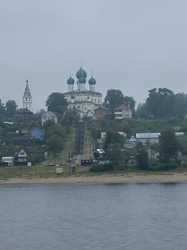 Высокий берег реки с деревянными домами, наверху белый собор с зелёно-синими куполами и отдельная колокольня, к воде спускается длинная лестница