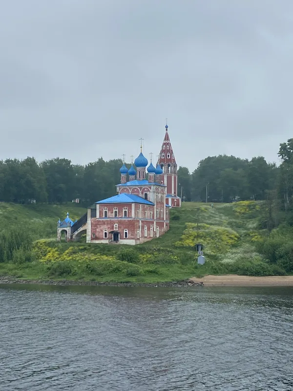 Краснокирпичная церковь с синими куполами и синей кровлей на зелёном берегу реки, рядом краснокирпичная шатровая колокольня