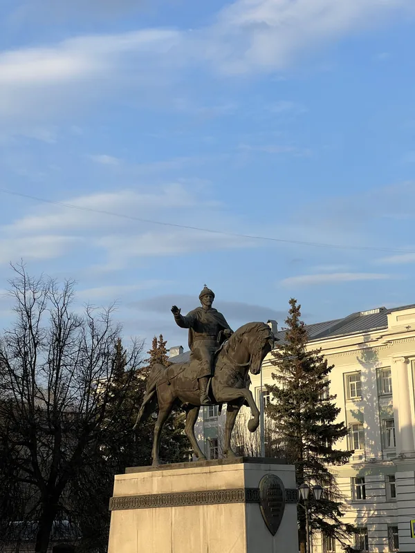 Бронзовый конный памятник на каменном постаменте: всадник в шлеме с поднятой рукой, за ним ели и светлое здание
