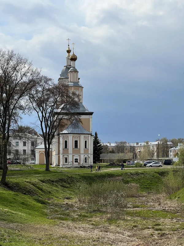 Светло-жёлтая церковь с золотой главкой и ярусной колокольней на краю зелёного поля, за ней невысокие городские дома