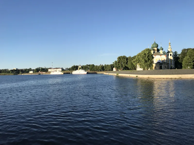 Широкая река в ясный день, справа на берегу церковь с золотыми куполами среди деревьев, вдалеке белые гидротехнические сооружения