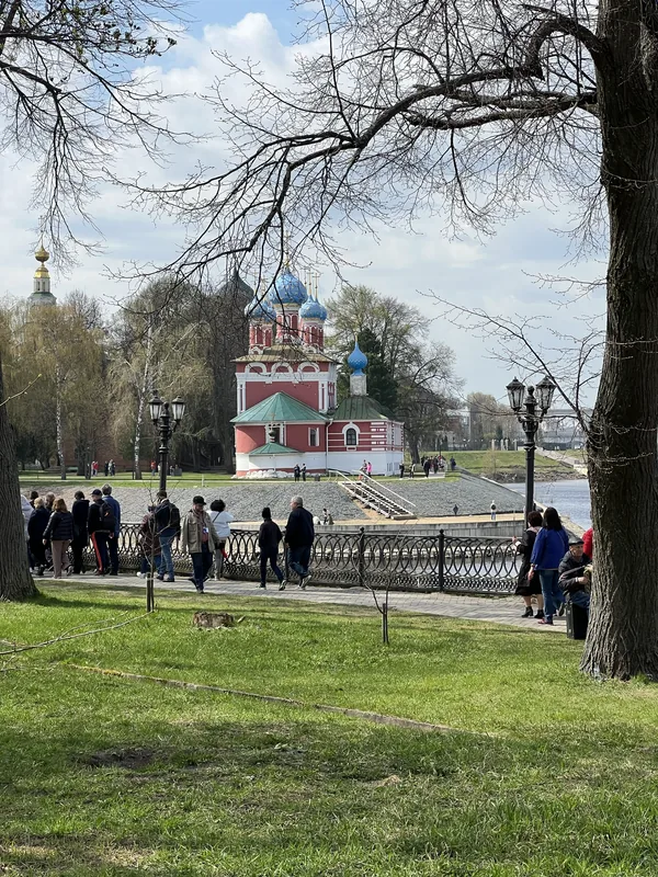 Краснокирпичная церковь с синими и золотыми куполами у реки, вдоль набережной с чугунной оградой гуляют люди, на переднем плане голые ветки дерева