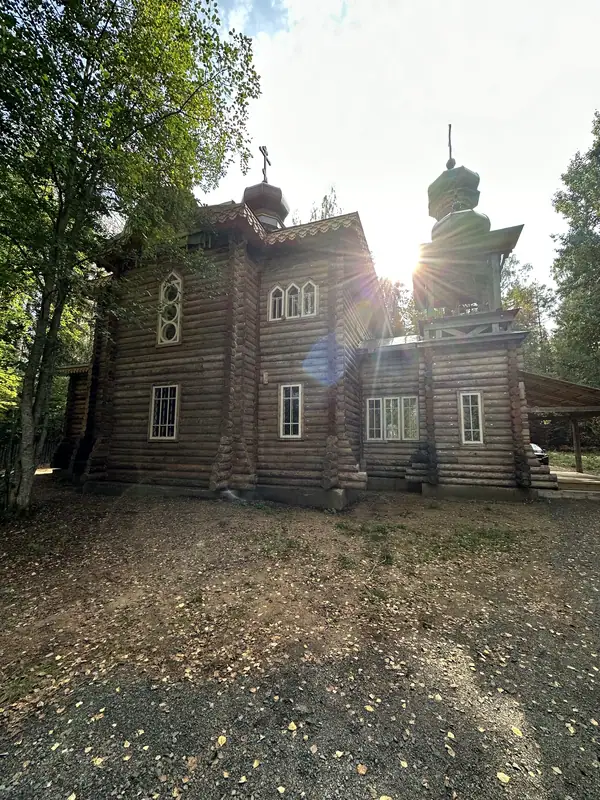 Бревенчатый храм с зелёными главами в лесу, солнце светит из-за колокольни
