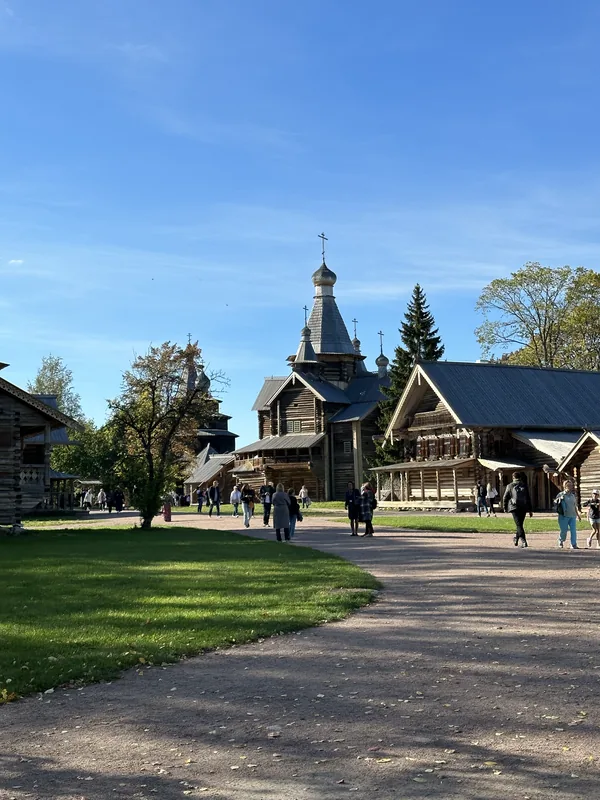 Витославлицы: деревянная шатровая церковь и срубы вдоль широкой дорожки, по газону и тропинкам гуляют посетители