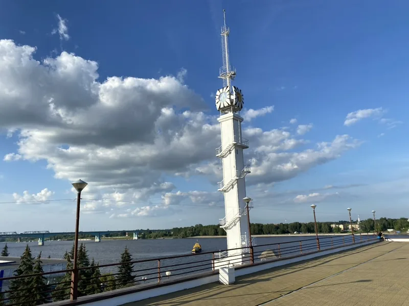 Высокая белая стела с часами и шпилем на набережной, за ней река с парусными лодками, вдоль парапета фонари