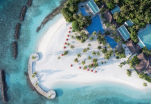 Мифы о Мальдивах, которые развеет Niva Dhigali Maldives Aerial view of a tropical beach resort with white sand, turquoise water, palm trees, and blue-roofed buildings along the shore.
