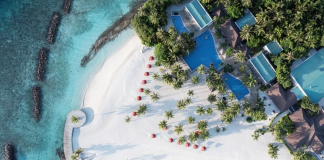 Aerial view of a tropical beach resort with white sand, turquoise water, palm trees, and blue-roofed buildings along the shore.