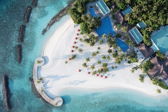 Aerial view of a tropical beach resort with white sand, turquoise water, palm trees, and blue-roofed buildings along the shore.