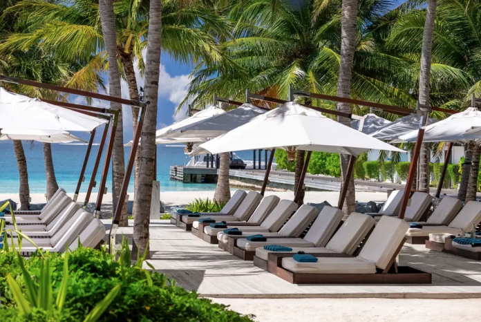 Снимок экрана 2026-05-19 в 19.18.27 Row of poolside lounge chairs under white umbrellas along a palm-lined beach with turquoise water in the background