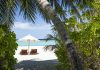 Two wooden lounge chairs under a white beach umbrella on a sunny tropical beach with turquoise water.