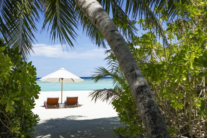 BeachLoungeSet-1-scaled Two wooden lounge chairs under a white beach umbrella on a sunny tropical beach with turquoise water.