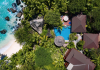 Aerial view of a tropical resort with turquoise sea, white beach, and dense palm trees surrounding brown-roofed villas and a blue pool.