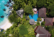 Обзор курорта Niva Labriz на Сейшелах Aerial view of a tropical resort with turquoise sea, white beach, and dense palm trees surrounding brown-roofed villas and a blue pool.