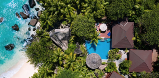 Aerial view of a tropical resort with turquoise sea, white beach, and dense palm trees surrounding brown-roofed villas and a blue pool.