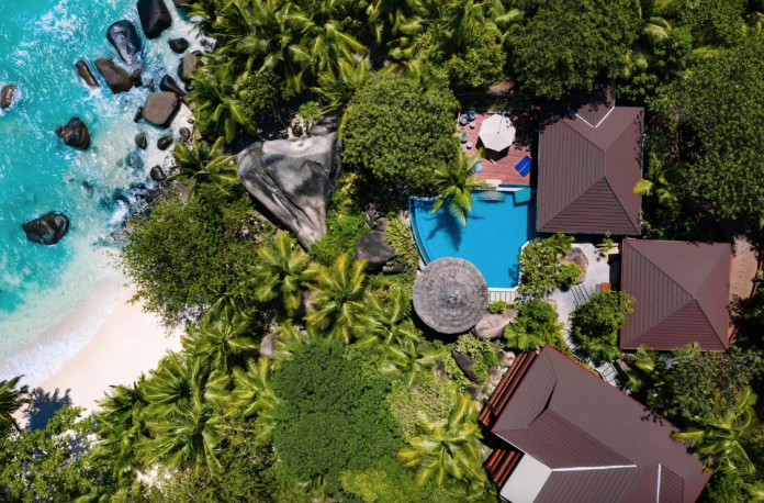Aerial view of a tropical resort with turquoise sea, white beach, and dense palm trees surrounding brown-roofed villas and a blue pool.