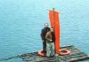 Two men stand on a weathered wooden raft with an orange sail/banner and a life preserver, drifting on calm blue water.