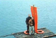Круизы для тех, кто никогда не был в круизе Two men stand on a weathered wooden raft with an orange sail/banner and a life preserver, drifting on calm blue water.