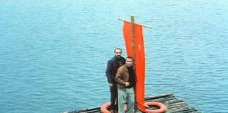 Two men stand on a weathered wooden raft with an orange sail/banner and a life preserver, drifting on calm blue water.